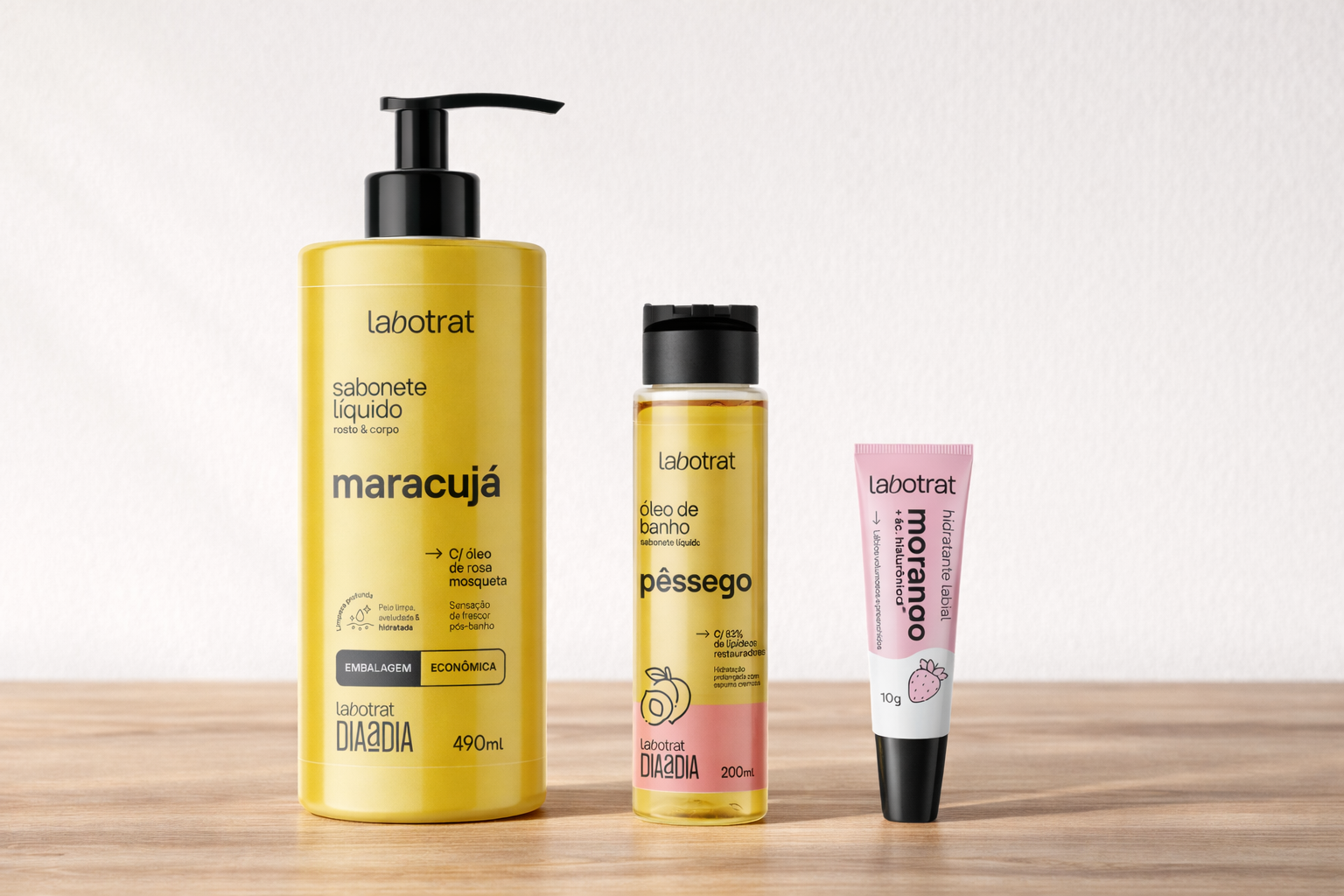 Produtos Labotrat dispostos sobre mesa de madeira com fundo branco, incluindo sabonete líquido Labotrat Maracujá 490ml com pump preto, óleo de banho Labotrat Pêssego 200ml em frasco transparente e hidratante labial Labotrat Morango 10g em bisnaga rosa e branca, embalagens originais preservadas e alinhadas em composição minimalista.
