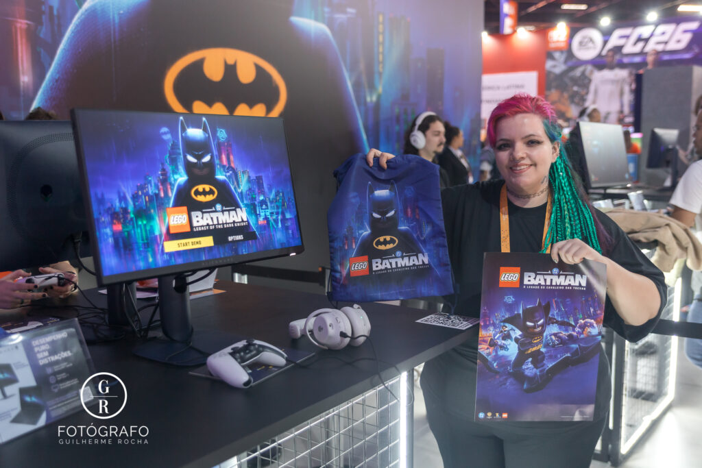 Visitante exibe brindes do jogo lego Batman, incluindo sacola e pôster, ao lado da estação de demo jogável no estande