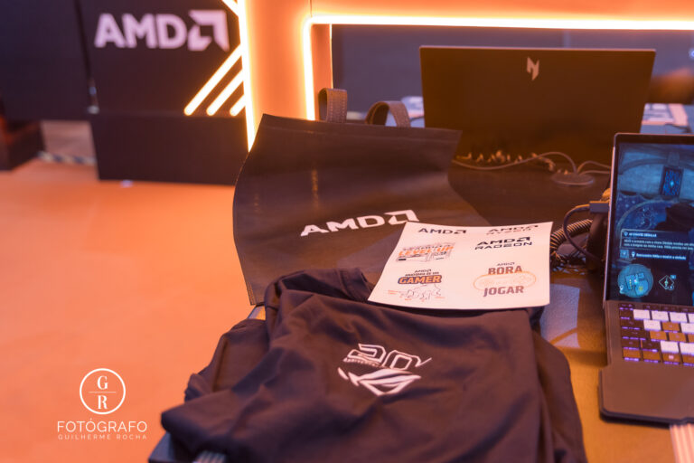 Brindes da AMD com camiseta, ecobag e adesivos distribuídos no estande com setup gamer ao fundo.