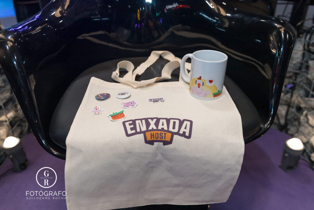 Ecobag enxada host exibida com brindes exclusivos como bottons, figurinhas e caneca durante evento gamer