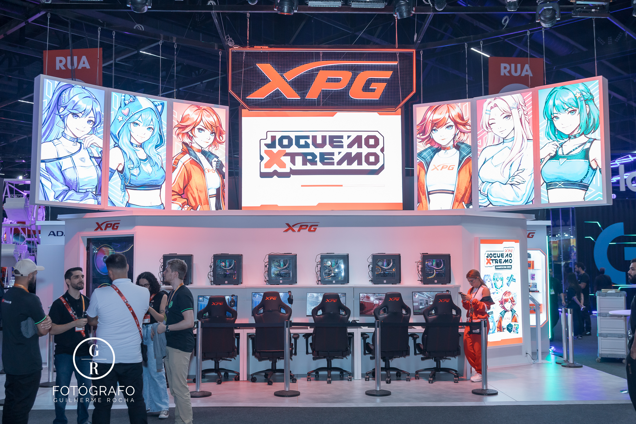 Estande da XPG na gamescom com área de free play equipada com vários PCs, cadeiras gamer e iluminação moderna, além de telões com personagens estilizados e branding da marca em destaque.