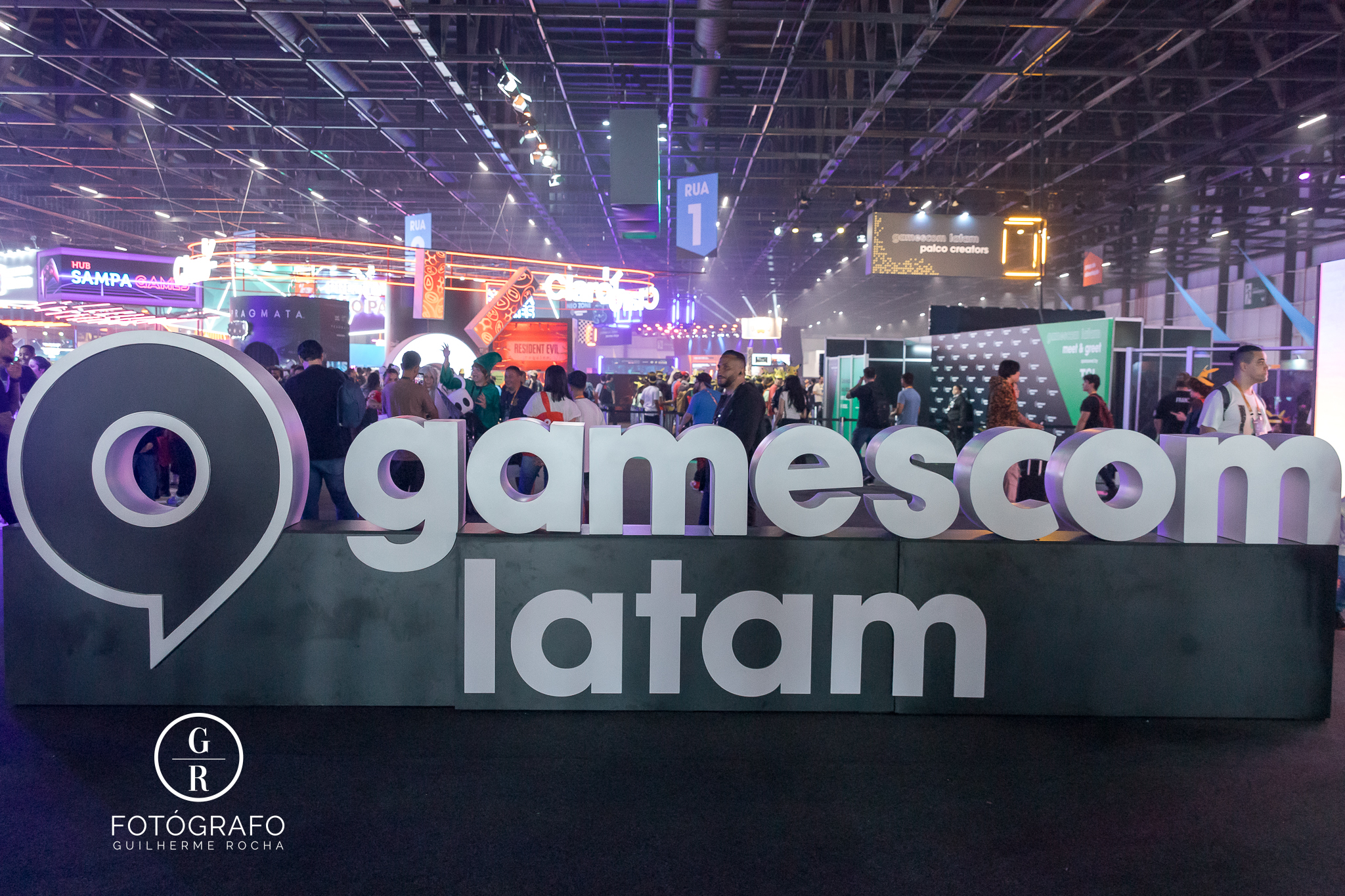 Vista do evento gamescom latam com grande letreiro em destaque no centro do pavilhão, rodeado por estandes iluminados, público circulando e atrações gamers ao fundo.