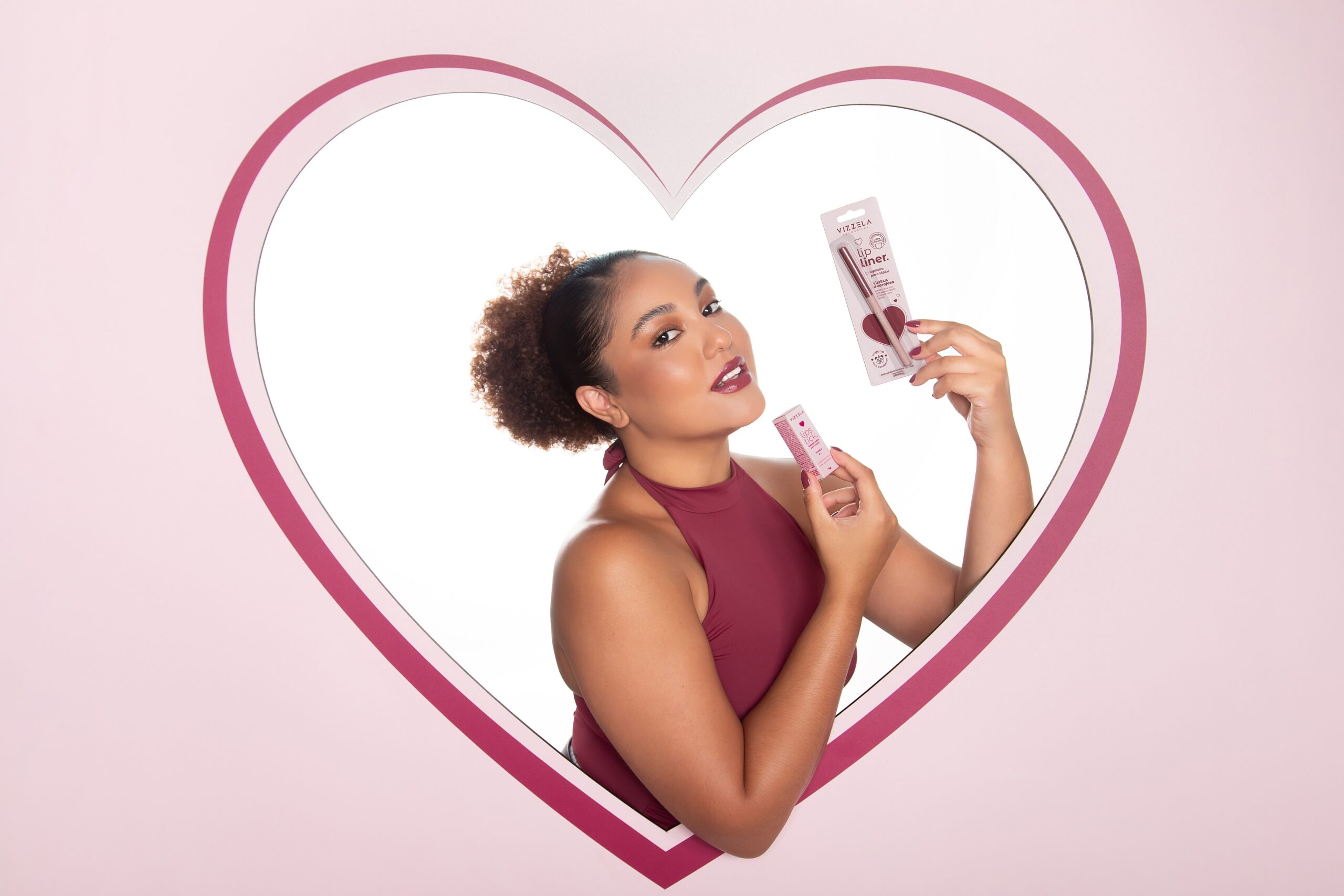 Modelo com maquiagem sofisticada segura batom e lapiseira labial vizzela dentro de moldura em formato de coração com fundo rosa suave.