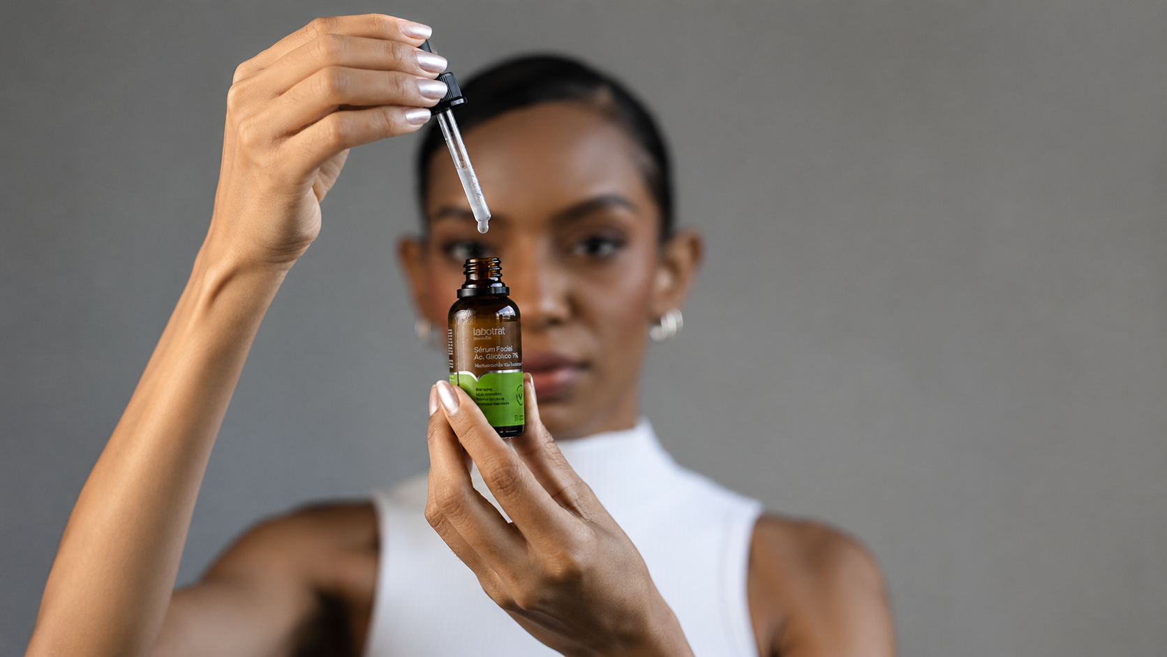 Sérum Facial Ácido Glicólico 7% sendo aplicado com conta-gotas por uma mulher. A imagem destaca o produto de skincare da Labotrat, focado em renovação celular, uniformização da pele e cuidados faciais. O frasco aparece em primeiro plano, enquanto a modelo surge desfocada ao fundo, reforçando a proposta de tratamento dermatológico e autocuidado.