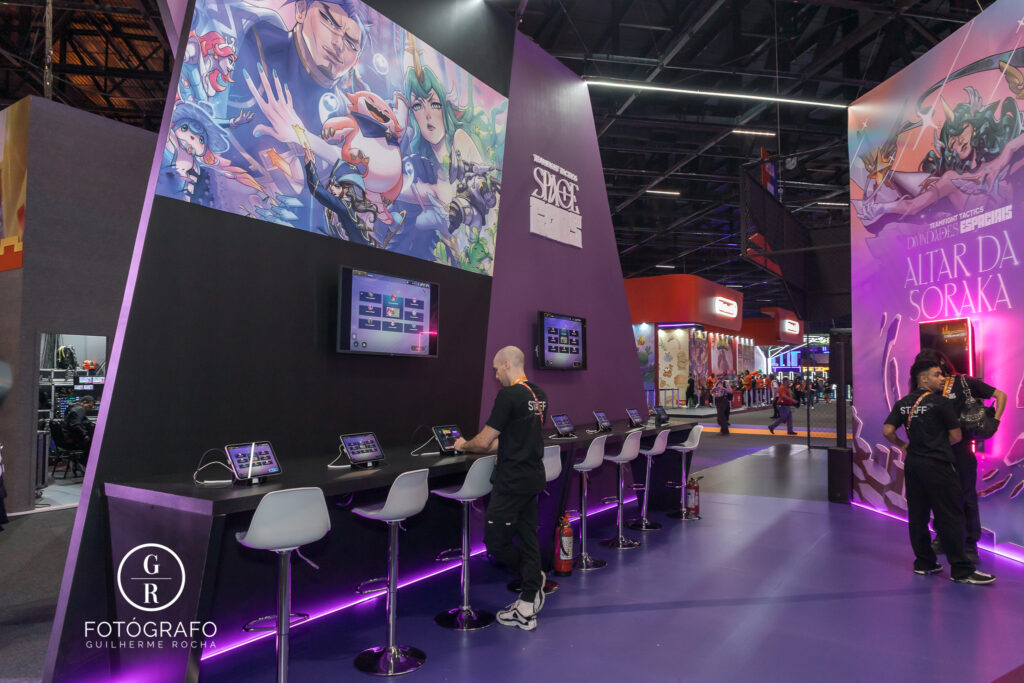 Estande da riot games com área de gameplay de TFT e telas interativas durante evento com iluminação roxa e temática de Runeterra
