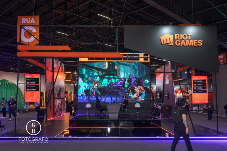 Estande principal da riot games com palco central, telão exibindo gameplay e programação visível ao público na gamescom