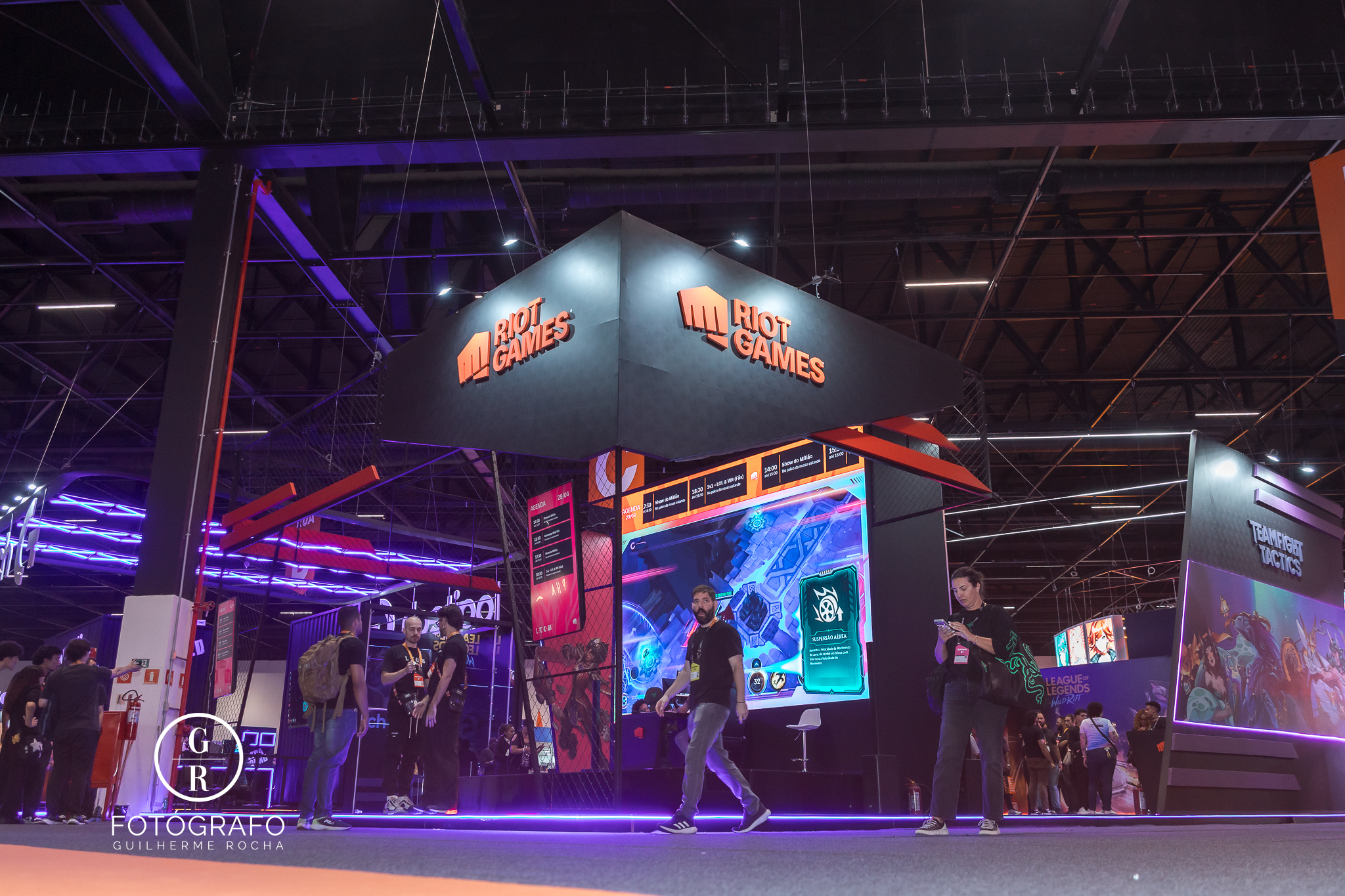 Visão lateral do estande da riot games com telões, área de jogos e visitantes circulando entre ativações do evento