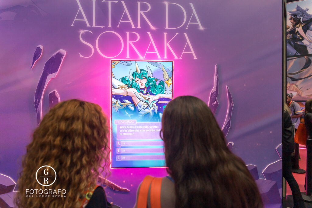 Visitantes participam do quiz Altar da Soraka no estande da riot games, interagindo com tela temática iluminada em rosa