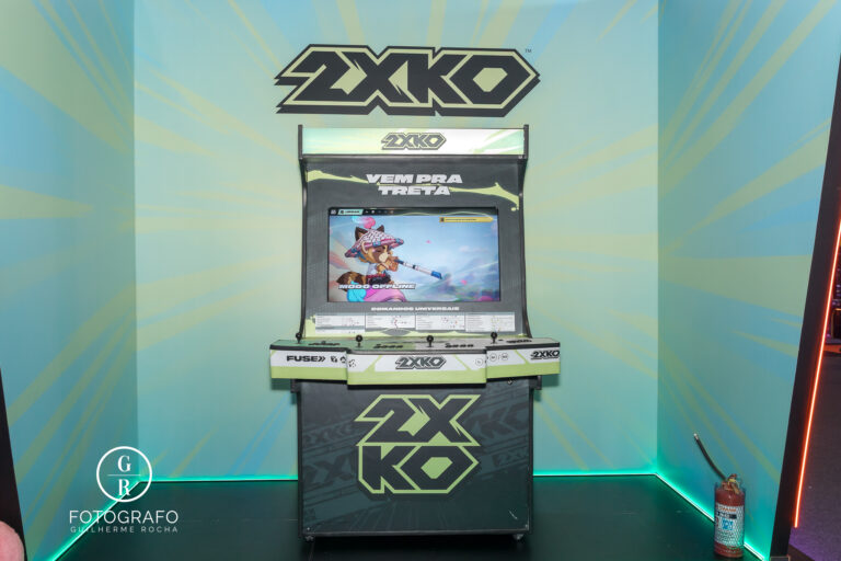 Máquina arcade temática em evento próxima ao estande da riot games com jogo exibido na tela e ambientação gamer colorida