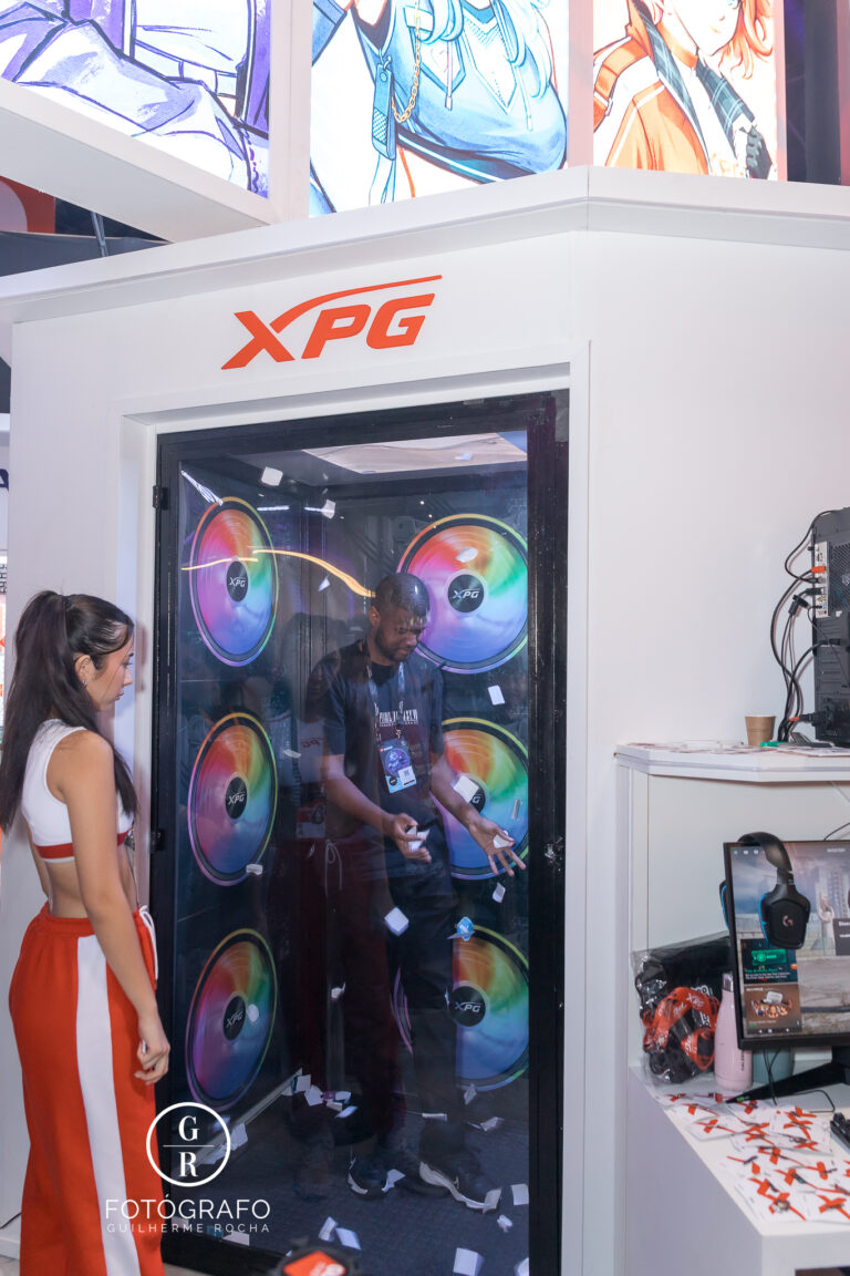 Cabine de vento da XPG com participante tentando pegar cupons enquanto papéis voam, durante dinâmica de brindes no estande.
