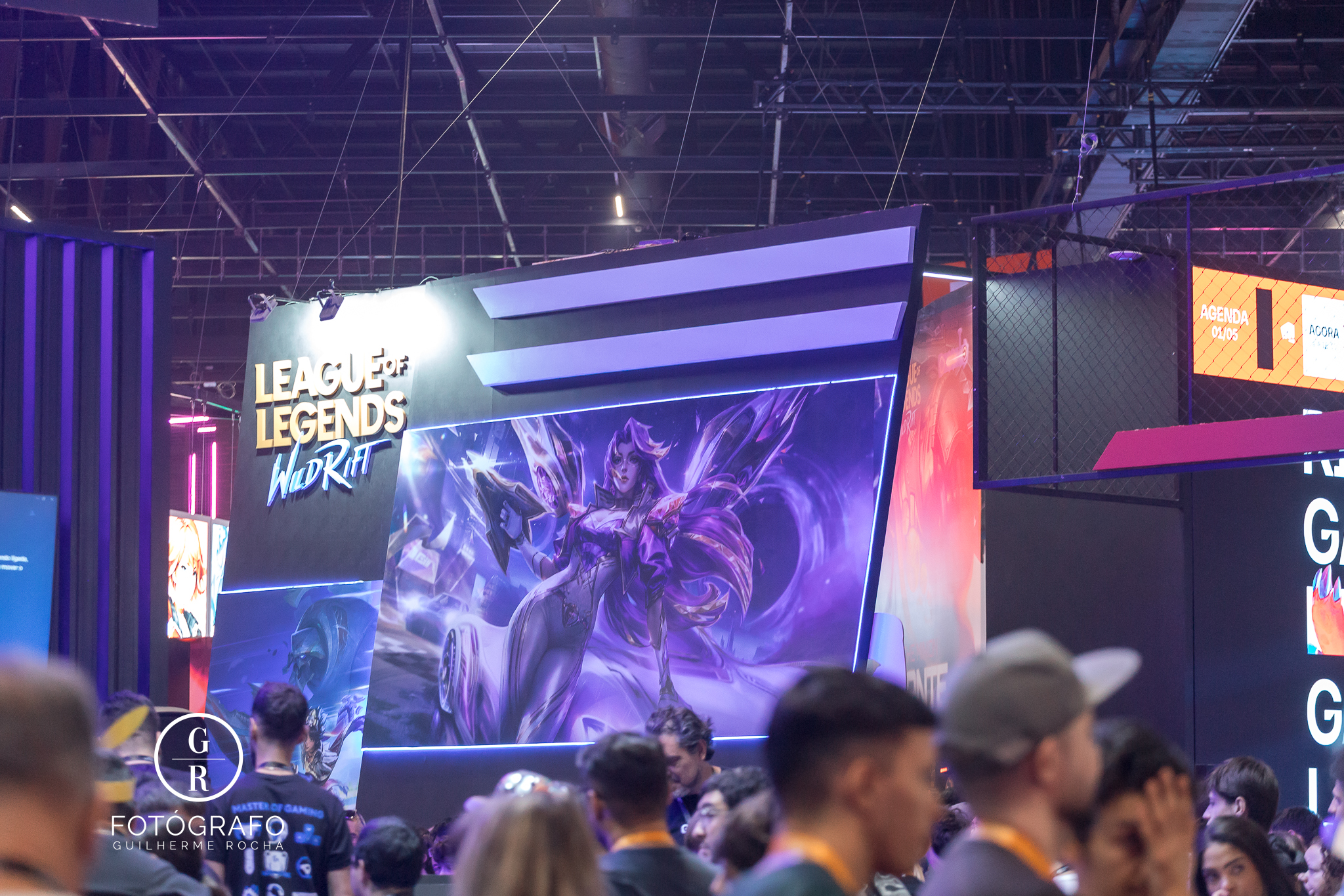 Estande de Riot Games com área dedicada a League of Legends: Wild Rift durante evento gamer, exibindo arte promocional da nova colaboração com Porsche enquanto visitantes circulam pelo espaço iluminado em tons neon.