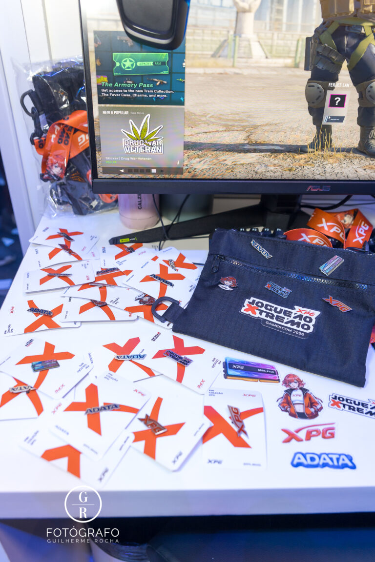 Mesa com brindes da XPG incluindo cartões, pins, adesivos e necessaire personalizada exibidos ao lado de estação de gameplay.
