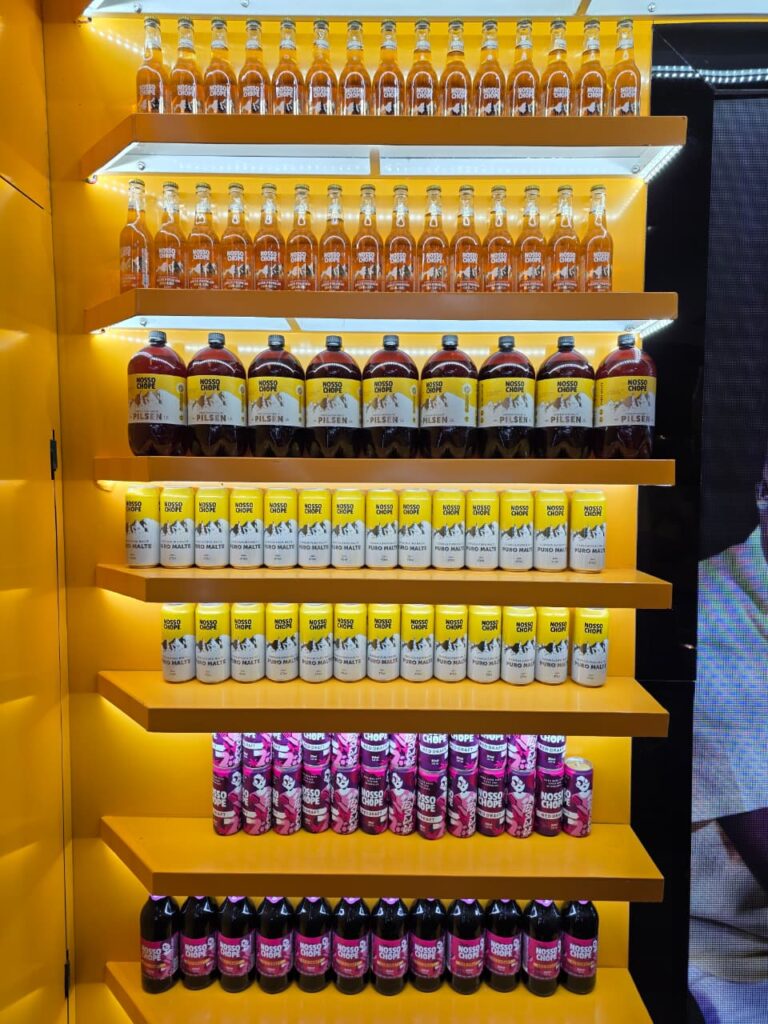 Prateleira iluminada do estande da Nosso Chope na APAS Show 2026 com cervejas puro malte, chope de vinho e embalagens variadas expostas ao público.