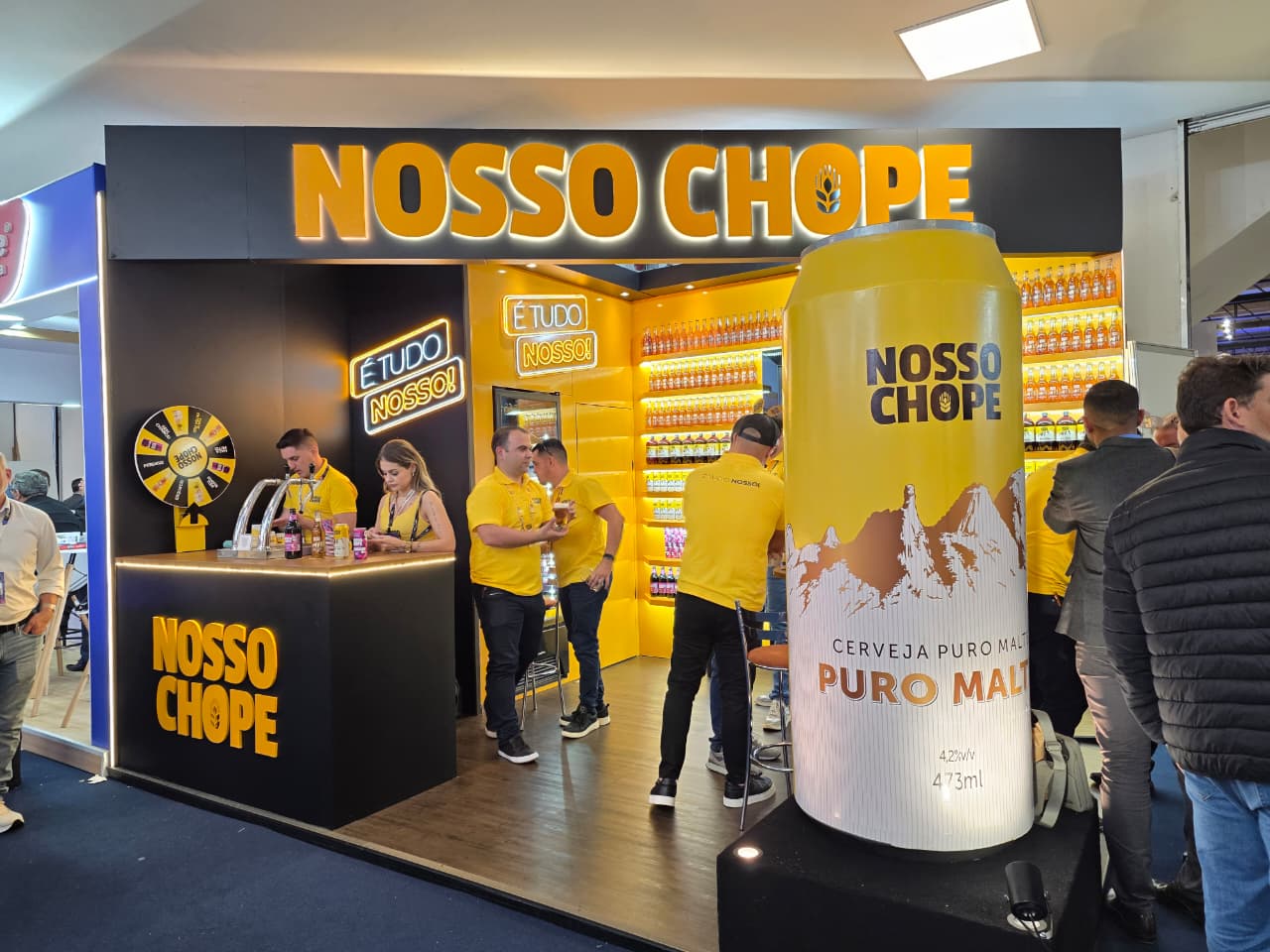 Estande da Nosso Chope na APAS Show 2026 com identidade visual iluminada, equipe de atendimento e destaque para a lata gigante de cerveja puro malte.