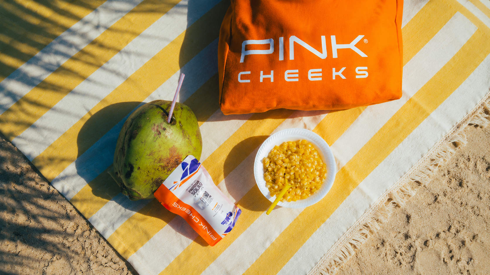 Produtos da PINK CHEEKS posicionados sobre toalha listrada na praia, com água de coco, snack e iluminação tropical vibrante.
