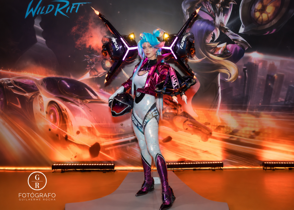 Cosplayer inspirada em personagem de Wild Rift posa durante ativação da Riot Games em evento gamer, com traje futurista neon, asas mecânicas iluminadas e cenário cyberpunk com carro esportivo ao fundo.
