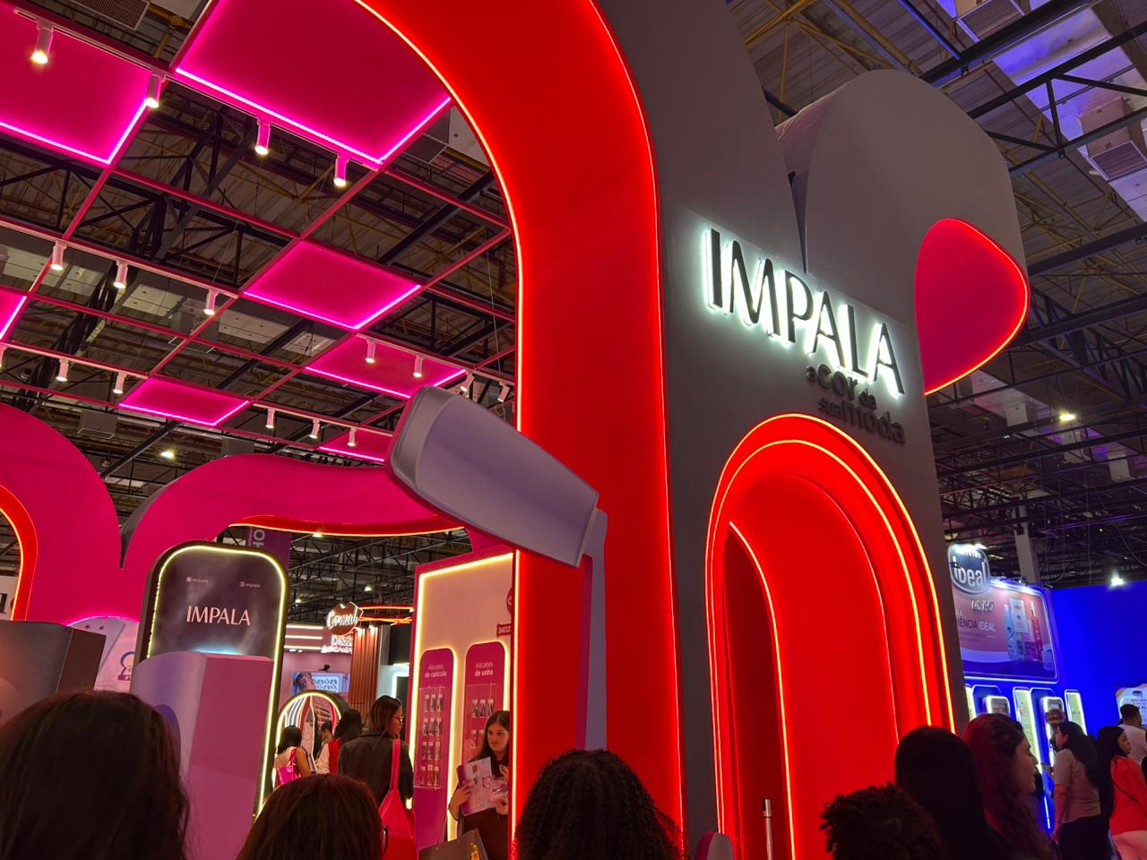 Estande da Impala na Beauty Show com iluminação rosa e vermelha, destacando a identidade visual da marca e o espaço dedicado aos lançamentos de esmaltes.