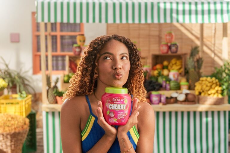 Mulher segurando um creme capilar da Skala em frente a uma banca colorida de frutas, destacando a energia da campanha Brazil Is Contagious e a conexão com a brasilidade.