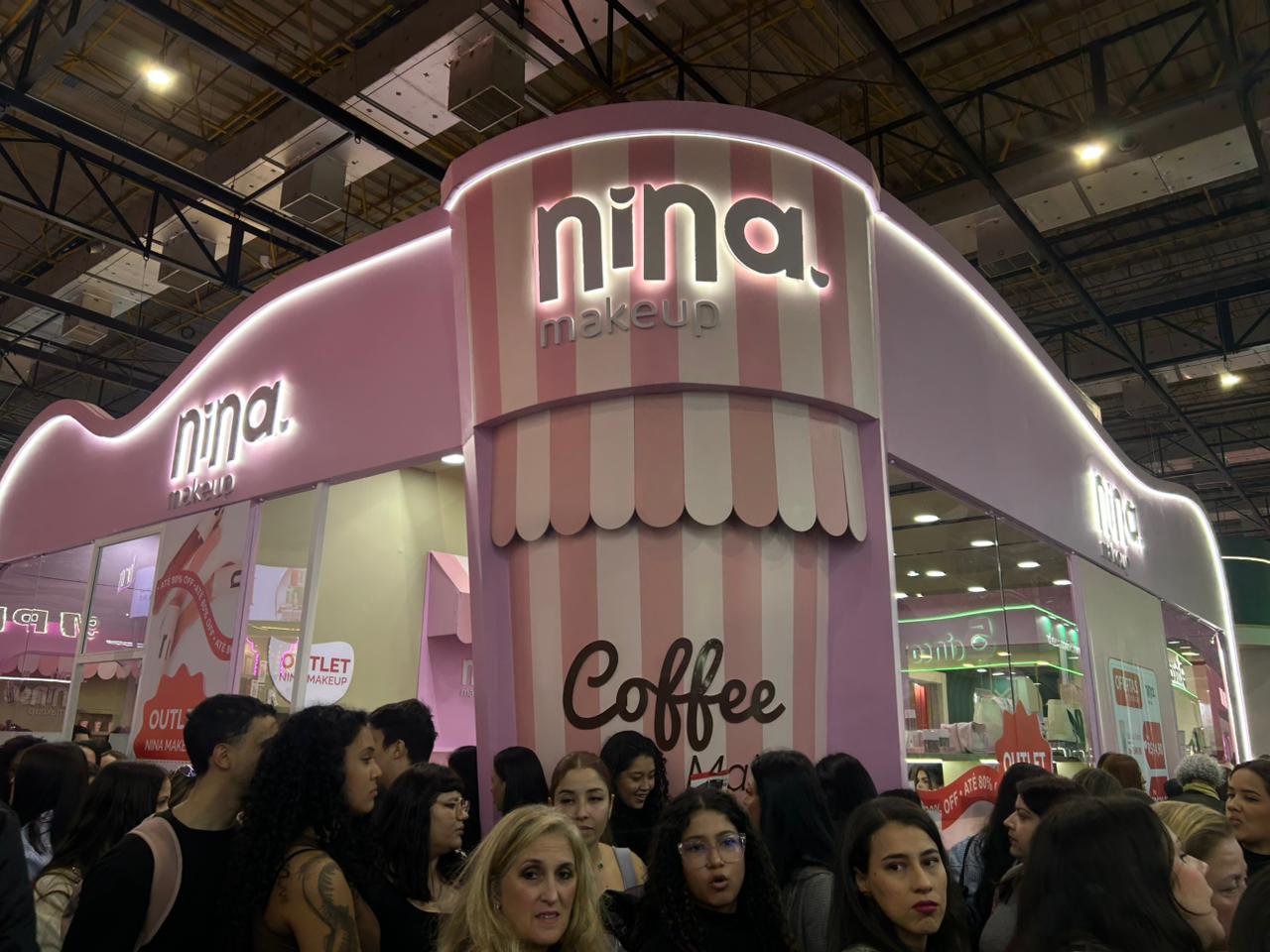 Estande da Nina Make Up na Beauty Show 2026 com fachada inspirada em cafeteria, identidade visual rosa e destaque para a linha Coffee Make, atraindo visitantes durante o evento.