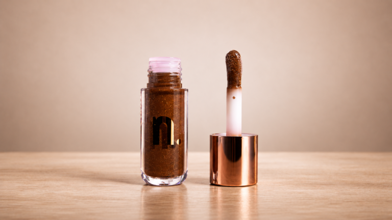 Nina Make Up Coffee Make Coffee: gloss labial com partículas douradas visíveis, frasco transparente aberto e aplicador em destaque, evidenciando o brilho e a textura inspirados no universo do café.