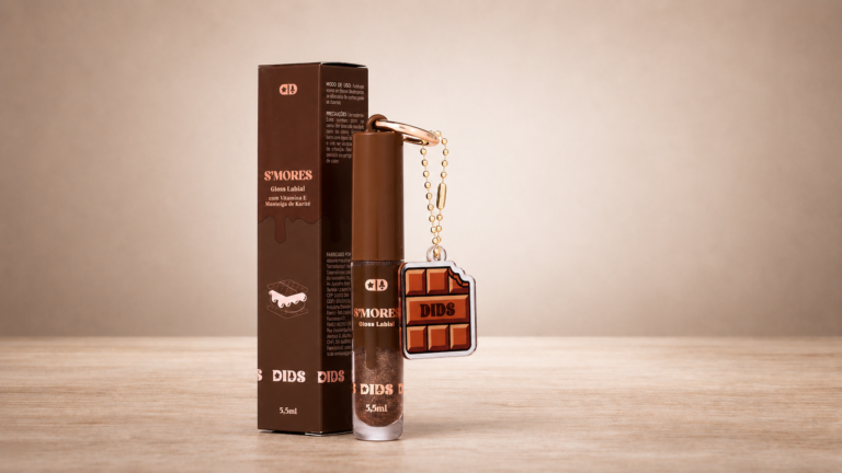 Dids Gloss Labial S'Mores com embalagem marrom inspirada em chocolate e chaveiro temático em formato de barra de chocolate. O produto aparece ao lado da caixa da coleção sobre superfície de madeira clara.
