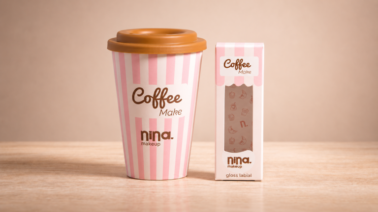 Nina Make Up Coffee Make: copo temático da coleção ao lado da embalagem do gloss labial, com design em listras rosa e brancas inspirado em cafeterias e edição limitada.