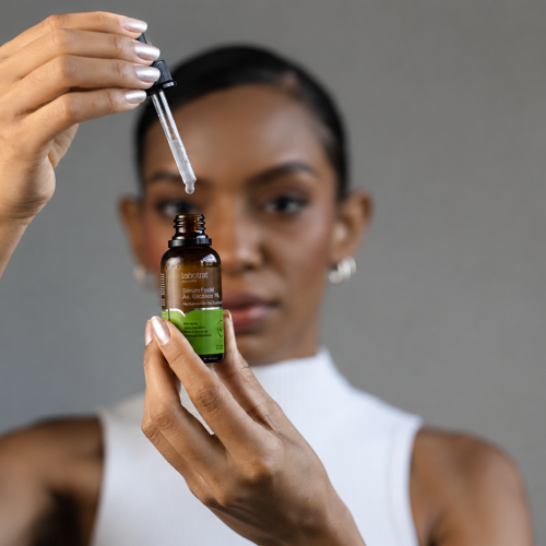 Sérum Facial Ácido Glicólico 7% sendo aplicado com conta-gotas por uma mulher. A imagem destaca o produto de skincare da Labotrat, focado em renovação celular, uniformização da pele e cuidados faciais. O frasco aparece em primeiro plano, enquanto a modelo surge desfocada ao fundo, reforçando a proposta de tratamento dermatológico e autocuidado.