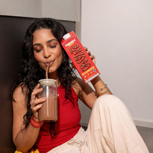 Mulher segura bebida gelada de chocolate vegetal enquanto bebe com canudo, ao lado de embalagem vermelha da Possible, em momento de consumo refrescante.