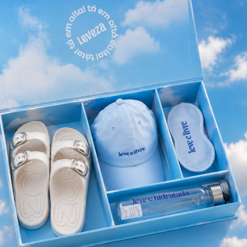 Kit recebido da Zaxy com sandália branca, boné, máscara de descanso e garrafa em caixa personalizada sobre fundo de céu azul com nuvens.