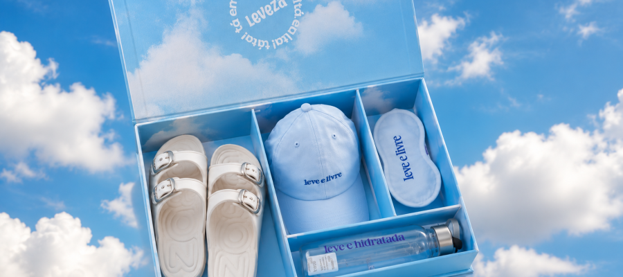 Kit recebido da Zaxy com sandália branca, boné, máscara de descanso e garrafa em caixa personalizada sobre fundo de céu azul com nuvens.