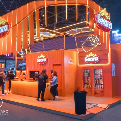 Estande da Seara na gamescom latam com iluminação laranja, área de atendimento e espaço para compra e consumo de produtos.