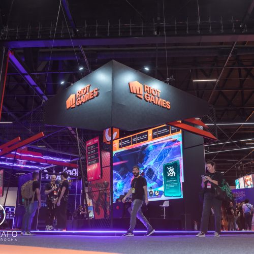 Visão lateral do estande da riot games com telões, área de jogos e visitantes circulando entre ativações do evento