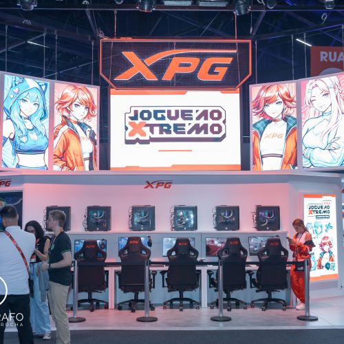 Estande da XPG na gamescom com área de free play equipada com vários PCs, cadeiras gamer e iluminação moderna, além de telões com personagens estilizados e branding da marca em destaque.