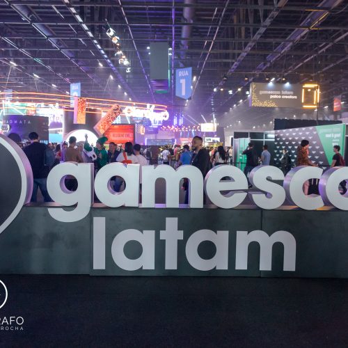 Vista do evento gamescom latam com grande letreiro em destaque no centro do pavilhão, rodeado por estandes iluminados, público circulando e atrações gamers ao fundo.