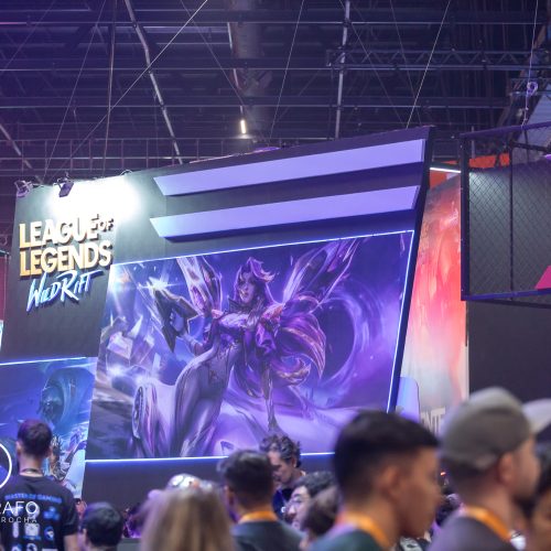 Estande de Riot Games com área dedicada a League of Legends: Wild Rift durante evento gamer, exibindo arte promocional da nova colaboração com Porsche enquanto visitantes circulam pelo espaço iluminado em tons neon.