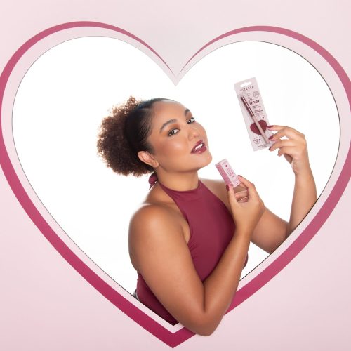 Modelo com maquiagem sofisticada segura batom e lapiseira labial vizzela dentro de moldura em formato de coração com fundo rosa suave.