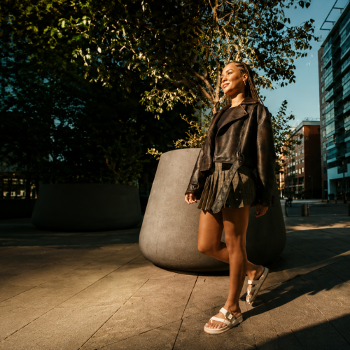 Modelo usa sandália Zaxy em produção de outono-inverno com jaqueta de couro e saia plissada, mostrando a versatilidade do calçado em diferentes estações.