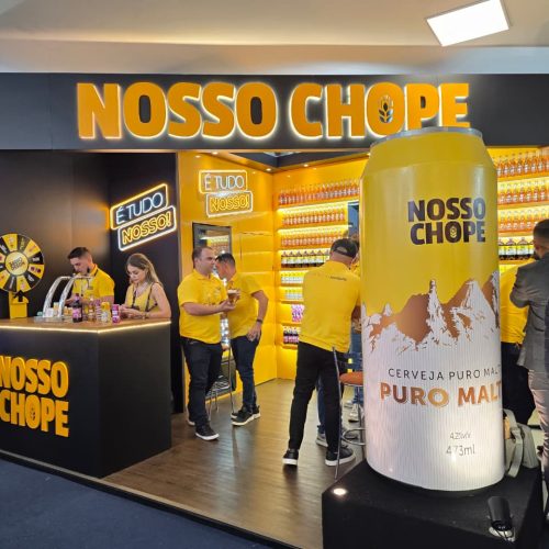 Estande da Nosso Chope na APAS Show 2026 com identidade visual iluminada, equipe de atendimento e destaque para a lata gigante de cerveja puro malte.