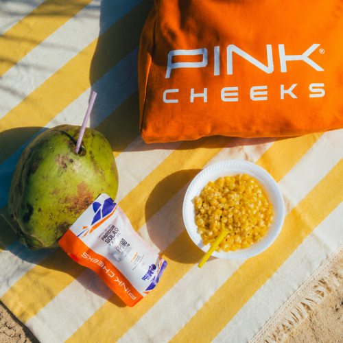 Produtos da PINK CHEEKS posicionados sobre toalha listrada na praia, com água de coco, snack e iluminação tropical vibrante.