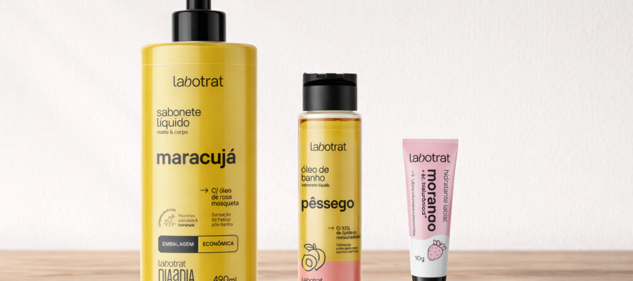 Produtos Labotrat dispostos sobre mesa de madeira com fundo branco, incluindo sabonete líquido Labotrat Maracujá 490ml com pump preto, óleo de banho Labotrat Pêssego 200ml em frasco transparente e hidratante labial Labotrat Morango 10g em bisnaga rosa e branca, embalagens originais preservadas e alinhadas em composição minimalista.